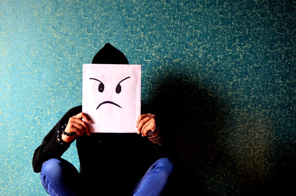 Person sitting on the floor, holding up a rectangular piece of paper or cardstock with an angry face painted on it. Mask.
Image by PDPics from Pixabay 