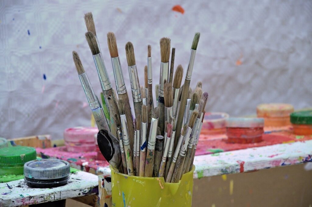 Container of paint brushes in the foreground. Behind it are paints and a white canvas. There are paint splashes everywhere. Artist studio.
Image by Jenny Shead from Pixabay 
