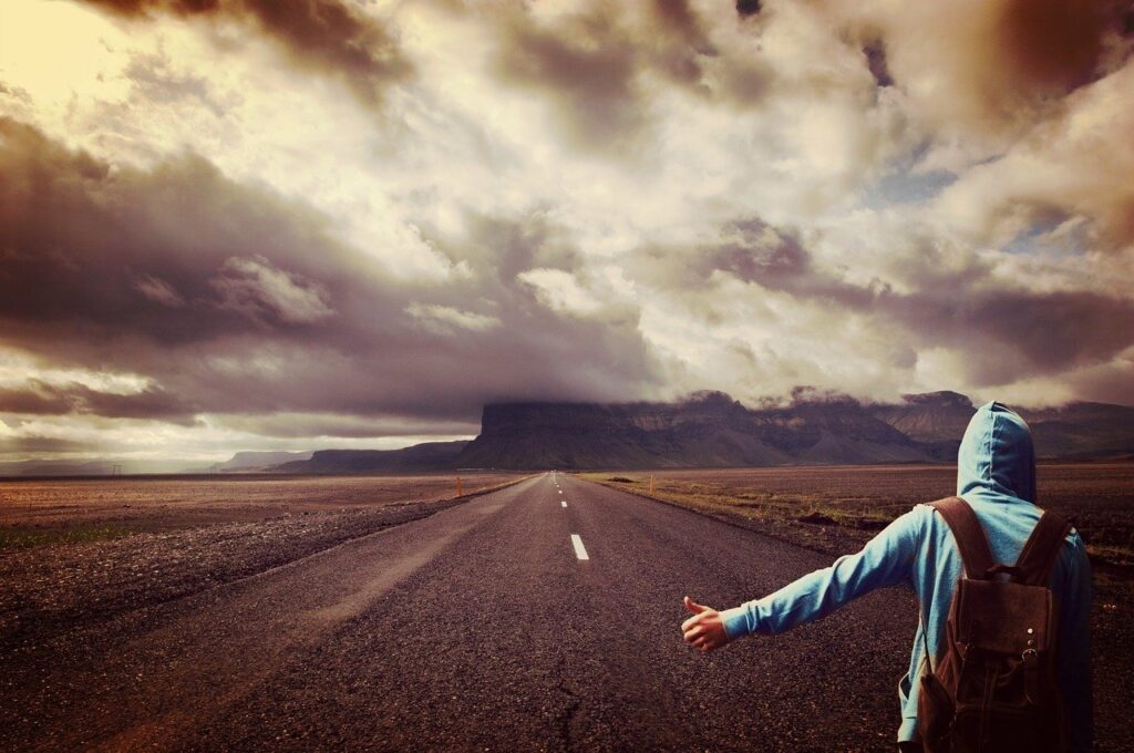 Photo of the back of a person wearing a hoodie and a backpack with his thumb out, hitchhiking down a two-lane highway. The road leads to low-lying mountains and dark clouds. The Prodigal Son. Lost. Lonely.
Image by cocoparisienne from Pixabay 