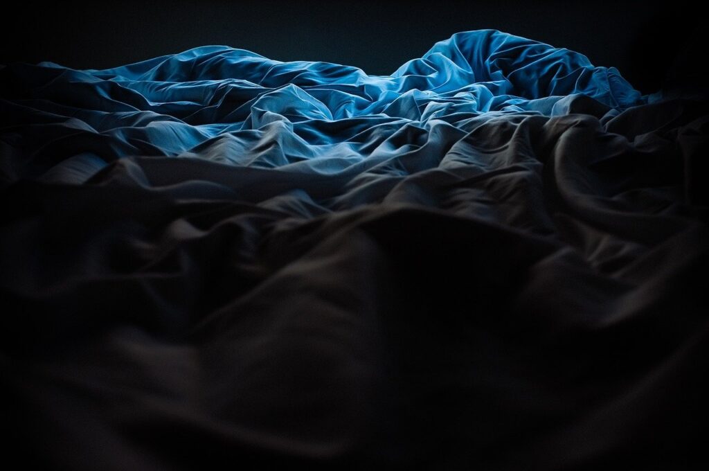 A blue comforter all rumpled. The end of it is highlighted by a light above, the bottom of the photo is in darkness so you can't make it out. Sleep, bed, sheets, sick bed
Image by Foundry Co from Pixabay 